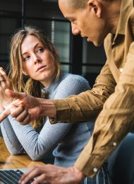 man in brown long sleeved button up shirt standing while using gray laptop computer on brown wooden table beside woman in gray long sleeved shirt sitting