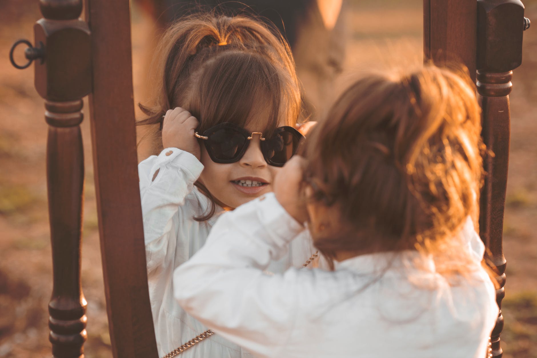 girl in white long sleeved shirt wearing sunglasses facing mirror